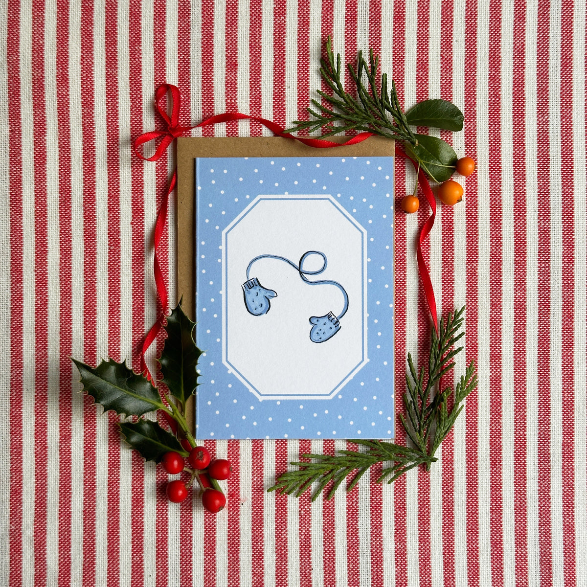 A mini A7 greeting card with a blue and white polka dot border and a central white octagonal panel showing a blue mitten and string illustration, styled on a red-and-white striped fabric with greenery and red ribbon.