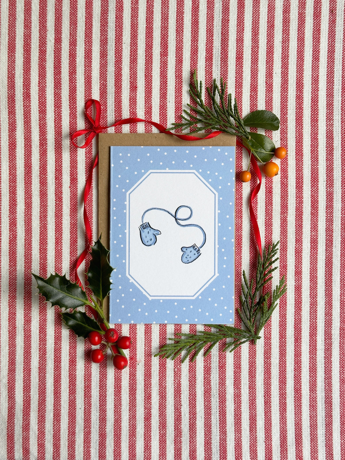 A mini greeting card with a blue and white design featuring two blue mittens, styled on a red and white striped fabric background with pine branches, holly leaves, and red berries.