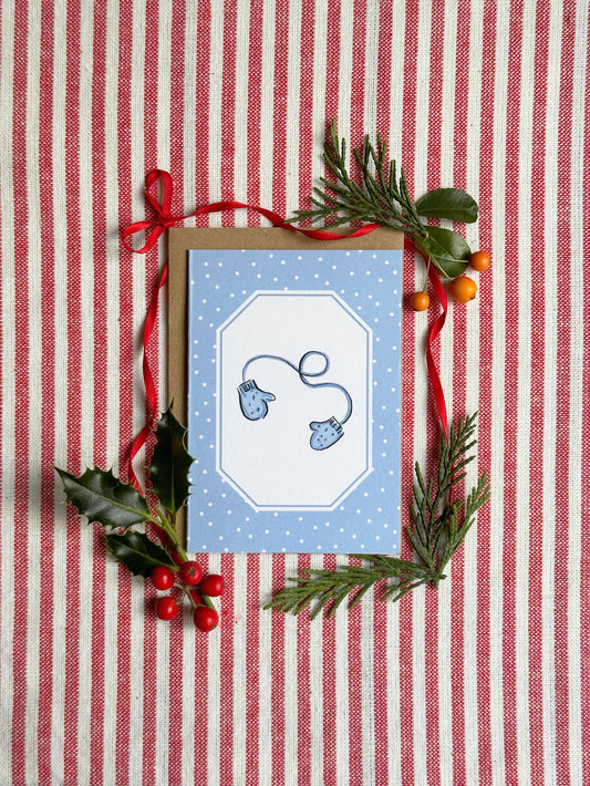 A mini greeting card with a blue and white design featuring two blue mittens, styled on a red and white striped fabric background with pine branches, holly leaves, and red berries.