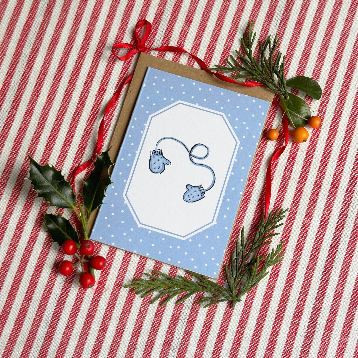 A mini A7 greeting card with a light blue border and white center, decorated with a blue mitten illustration, placed on a red-and-white striped fabric background with holly leaves, pine branches, and a red ribbon.