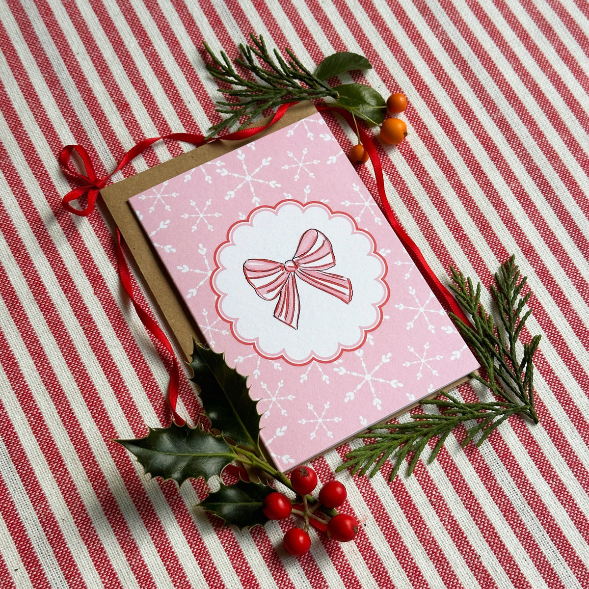 A pink Christmas greeting card with a scalloped white border and a bow illustration, placed on a brown envelope over a red-and-white striped fabric. The card is styled with green pine sprigs, holly leaves, and red berries.