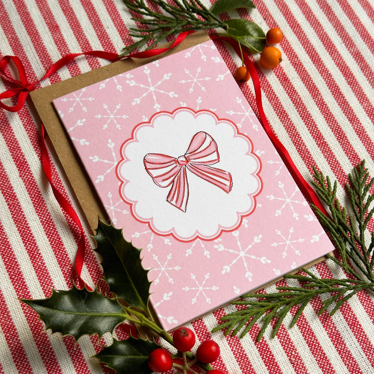 A pink Christmas greeting card with a scalloped white border and a red-and-white striped bow illustration, placed on a red-and-white striped fabric with green holly leaves, pine branches, and red berries.