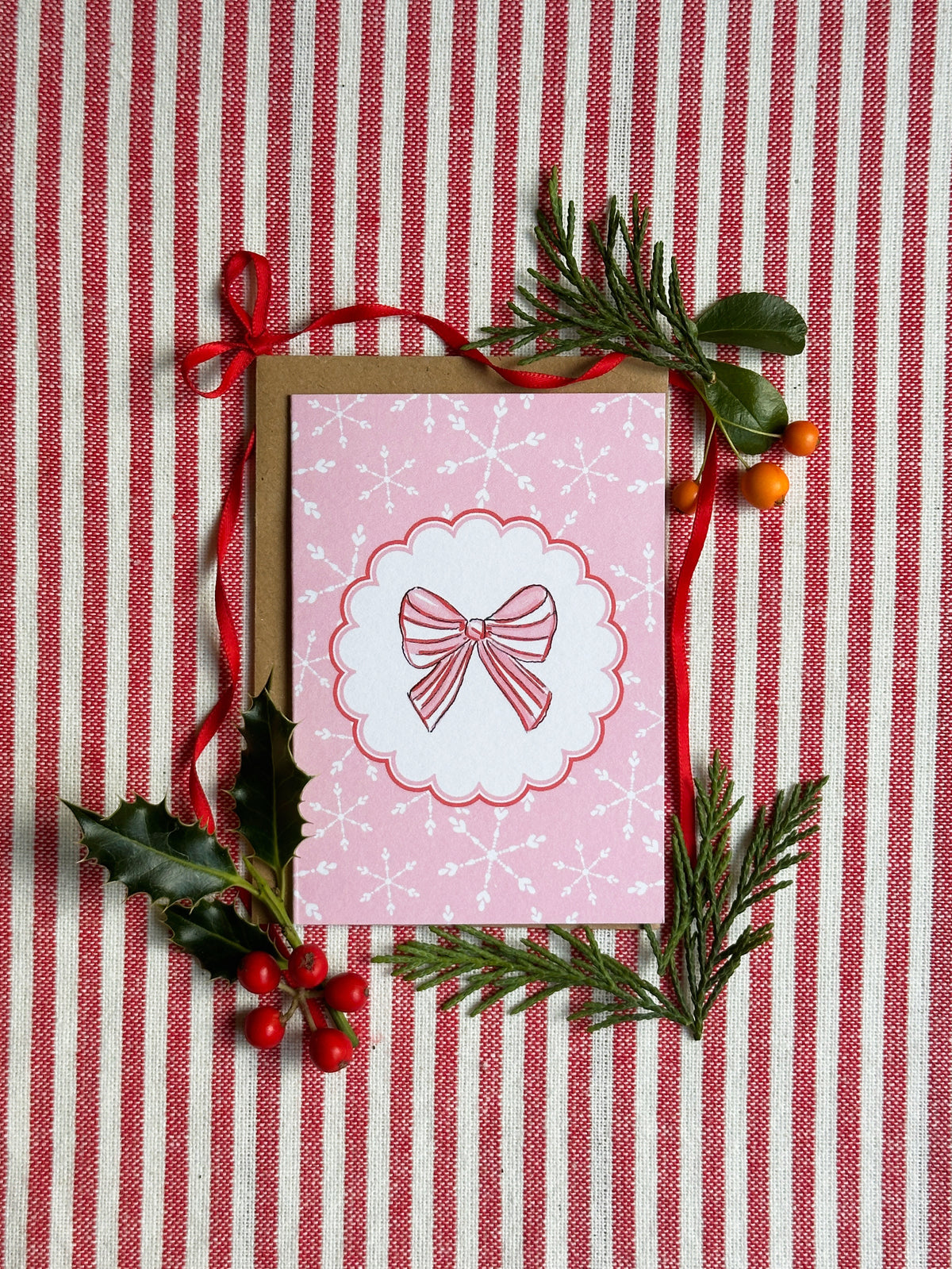 A mini a7  pink Christmas greeting card with a white bow illustration, styled on a red and white striped fabric background with pine branches, holly leaves, and red berries.