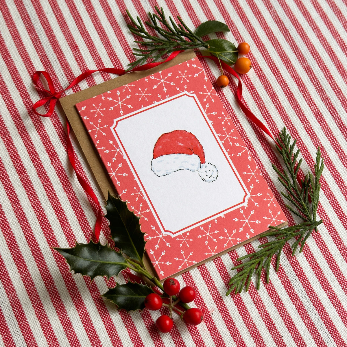 A Christmas greeting card with a red border, white snowflake pattern, and a Santa hat illustration, styled on a red and white striped fabric with green pine branches, holly leaves, and red ribbon.
