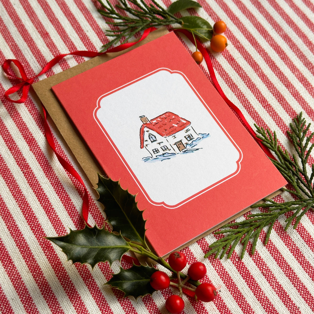 A red greeting card with a white central label showing a small illustrated house, placed on a brown envelope over a red-and-white striped fabric background with holiday greenery and red berries.