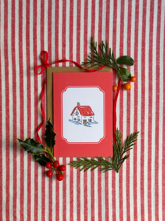 A red Christmas greeting card with a watercolor illustration of a cozy house, styled with a kraft envelope, red ribbon, holly leaves, and pine branches on a red and white striped fabric background.