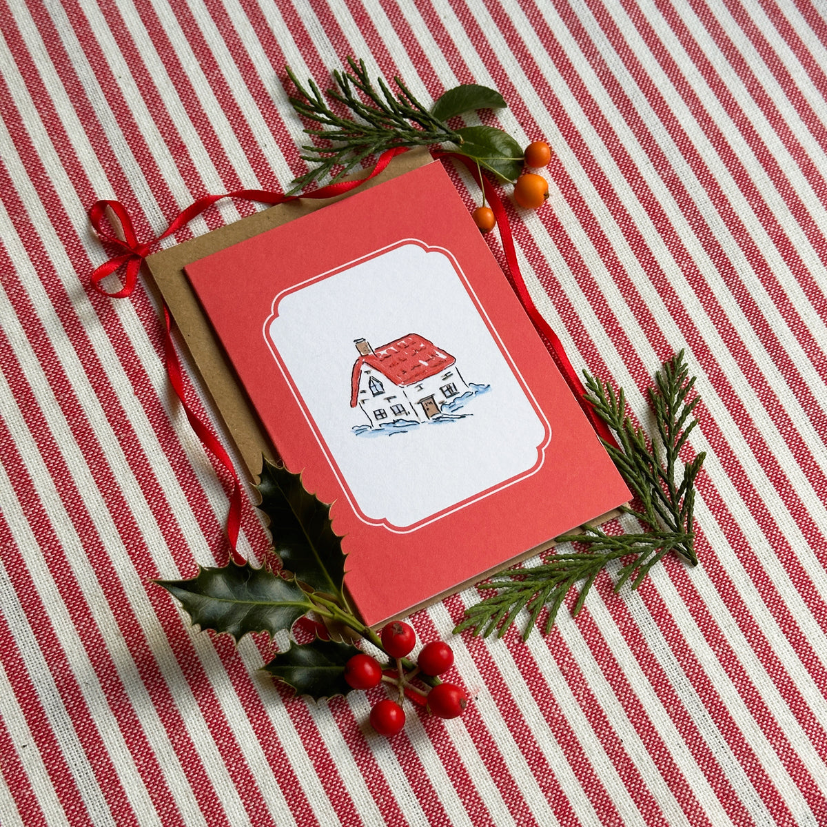 A red Christmas greeting card with a white central panel showing a small house illustration, placed on a kraft envelope and arranged on a red-and-white striped fabric with evergreen sprigs and red berries.
