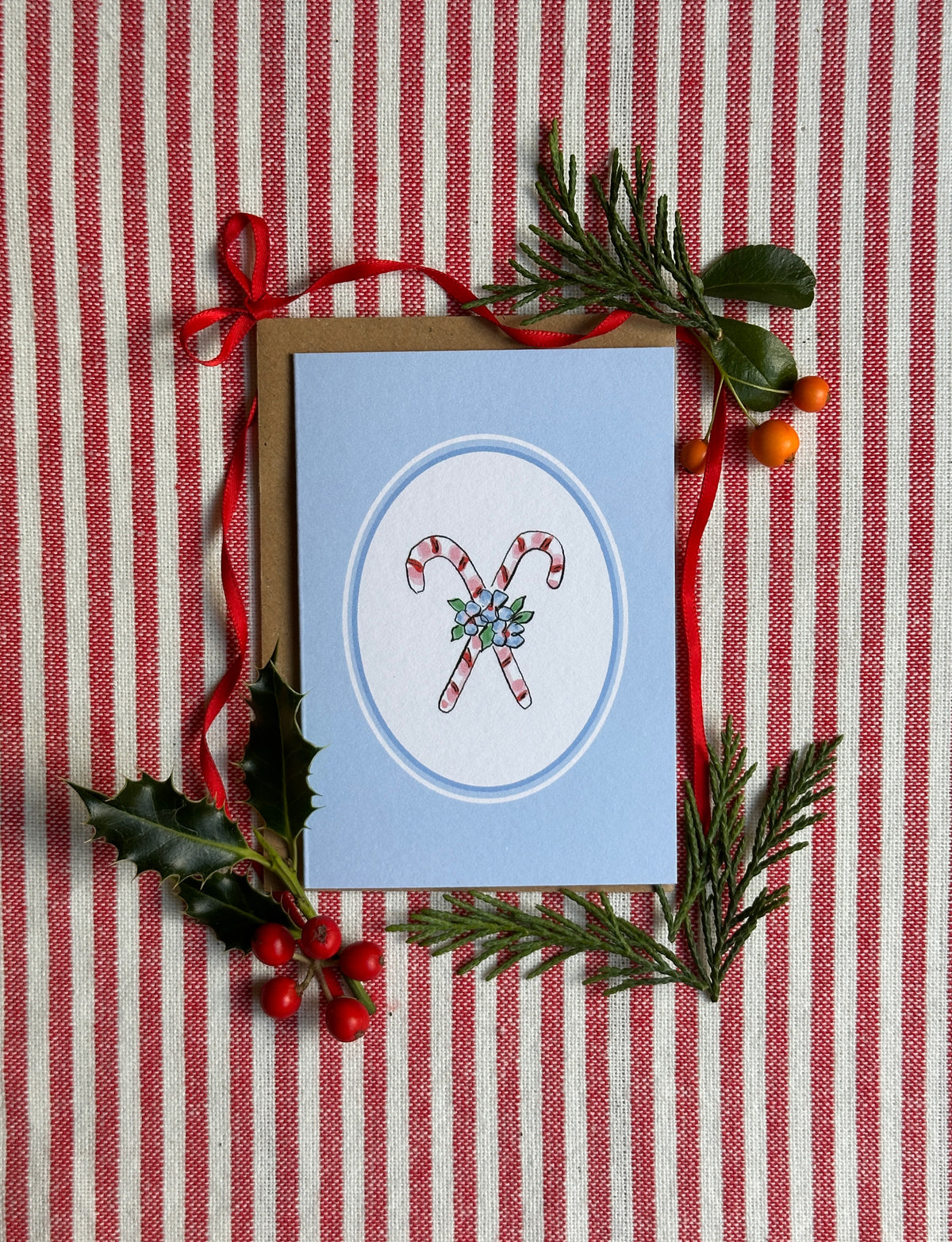 A mini Christmas greeting card with a light blue cover, featuring an illustration of two candy canes in a circular frame. The card is displayed on a red and white striped fabric background and styled with a brown envelope, red ribbon, holly leaves, pine branches, and small red berries.
