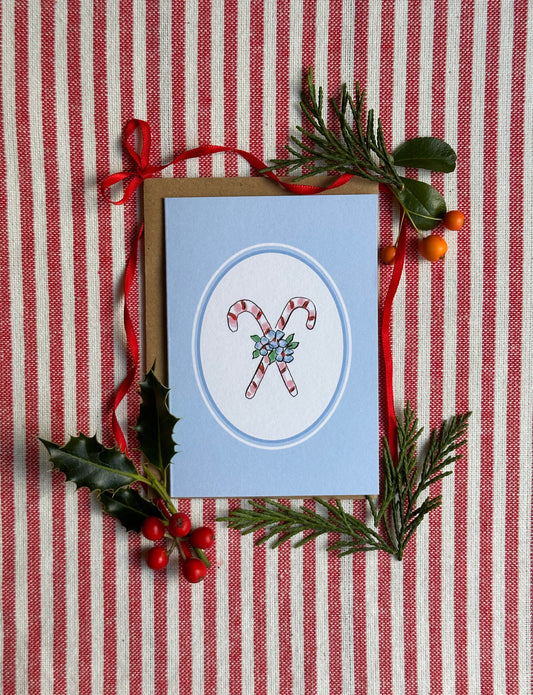A mini Christmas greeting card with a light blue cover, featuring an illustration of two candy canes in a circular frame. The card is displayed on a red and white striped fabric background and styled with a brown envelope, red ribbon, holly leaves, pine branches, and small red berries.