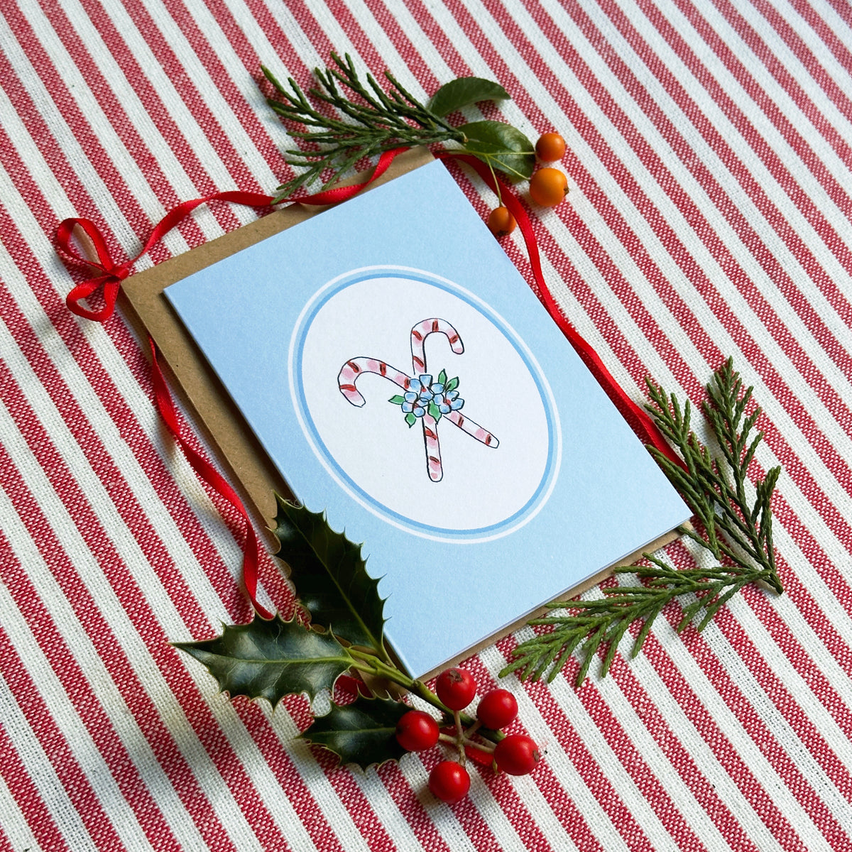 A light blue Christmas greeting card with a kraft envelope, styled on a red and white striped fabric background with holly leaves, pine branches, and red berries. The card features a white oval center with an illustration of two candy canes wrapped in a sprig of mistletoe.