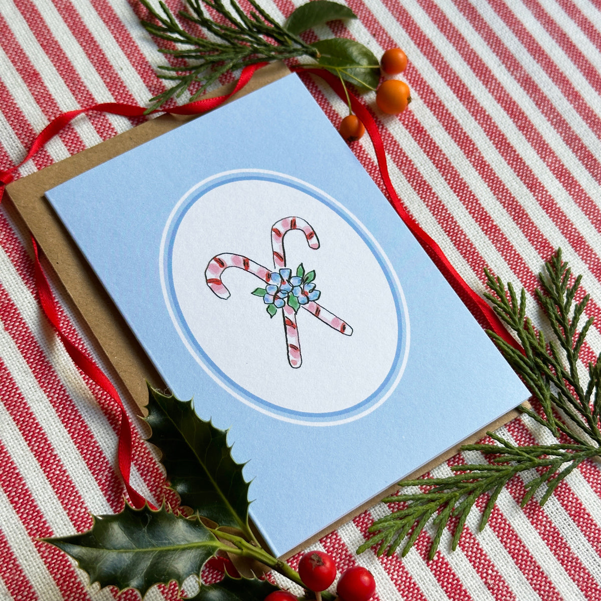 A light blue greeting card with a white oval center featuring two illustrated candy canes and small green/blue floral details, styled on a red-and-white striped fabric with evergreen branches, holly leaves, and red berries.