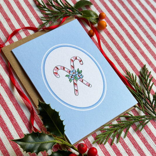 A light blue greeting card with a white oval center featuring two illustrated candy canes and small green/blue floral details, styled on a red-and-white striped fabric with evergreen branches, holly leaves, and red berries.