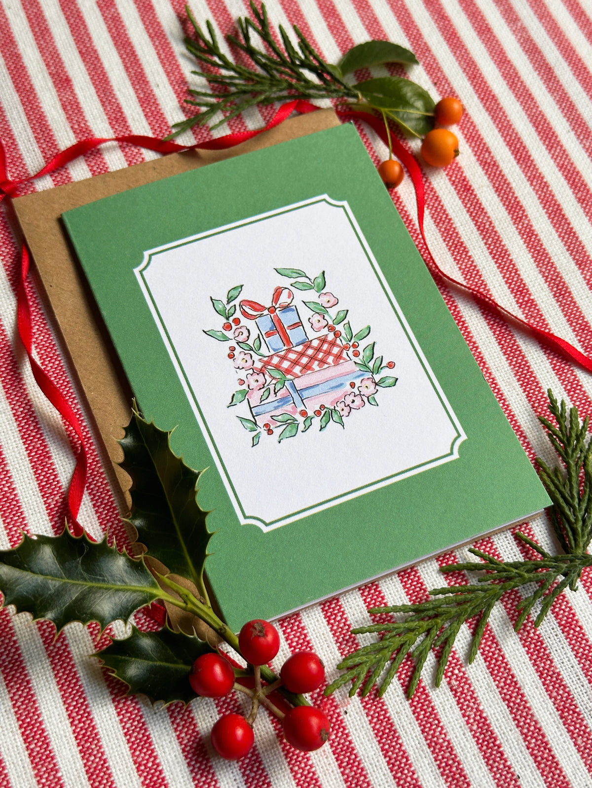 A green Christmas greeting card with a white border and an illustration of wrapped gifts, flowers, and a red bow, placed on a brown envelope over a red-and-white striped fabric with holly and pine branches.