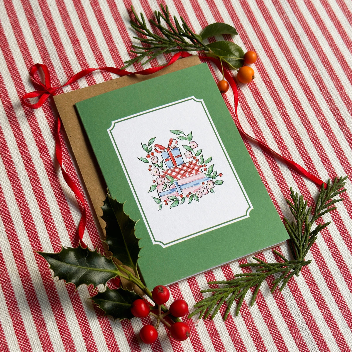 A green Christmas greeting card with a white central panel showing an illustration of a gift box, evergreen branches, holly leaves, and red berries, arranged on a red-and-white striped fabric with a red ribbon.