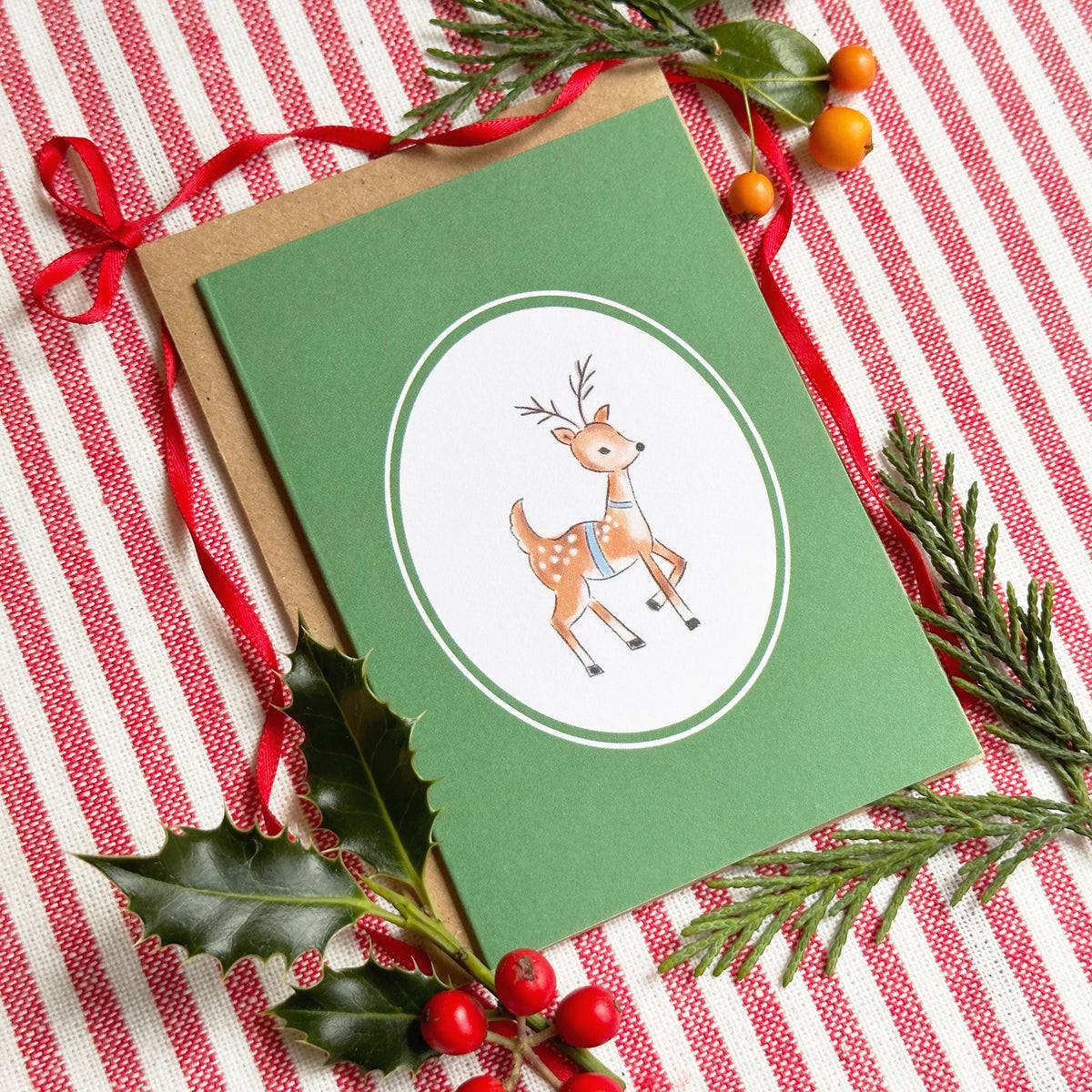 A green Christmas greeting card with a white oval featuring a brown deer illustration, styled on a red-and-white striped fabric with a kraft envelope, red ribbon, holly leaves, and pine branches.