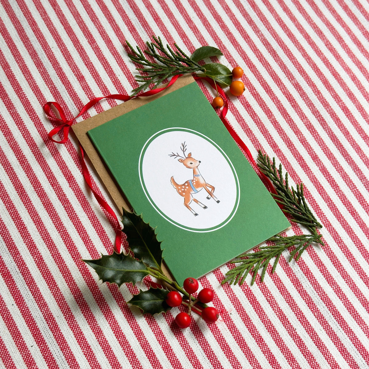 A green Christmas greeting card with a white oval featuring a cartoon deer, styled with a kraft envelope, red ribbon, holly leaves, and red berries on a red and white striped fabric background.
