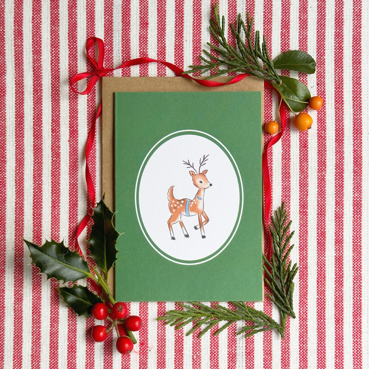 A green Christmas greeting card with a white oval containing a cute illustrated deer, styled on a red and white striped fabric with holly leaves, pine branches, and red berries.