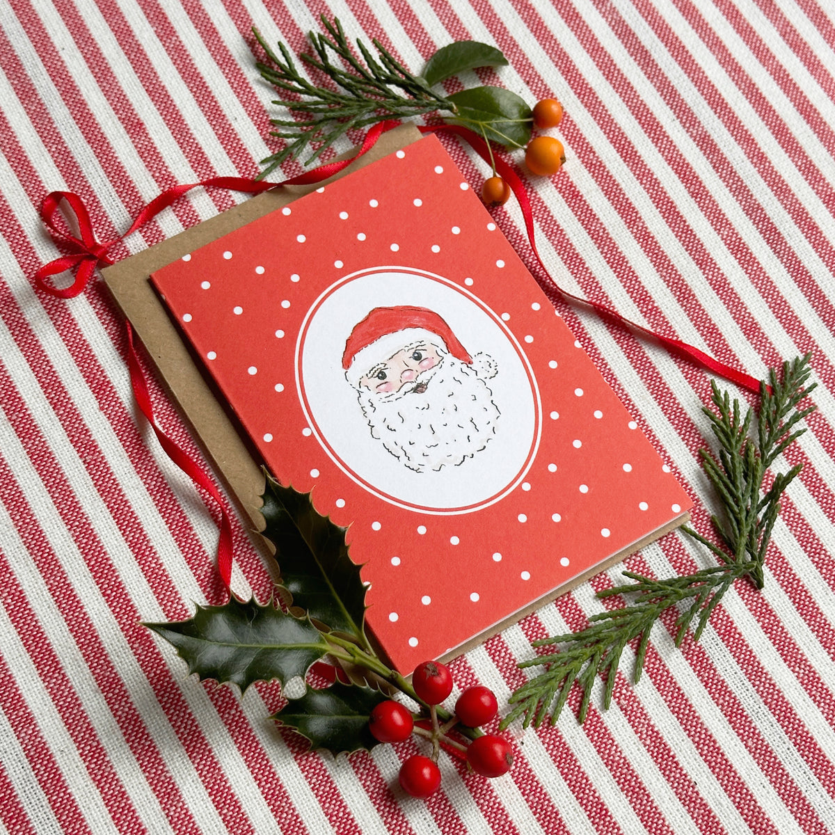 A red Christmas greeting card featuring a white oval with a cartoon Santa face, styled on a red and white striped fabric with pine branches, holly leaves, and red berries.