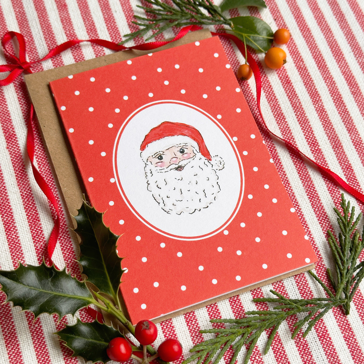 A red Christmas greeting card with white polka dots and an oval illustration of Santa's face, placed on a red-and-white striped fabric with holly leaves, pine branches, and a brown envelope.