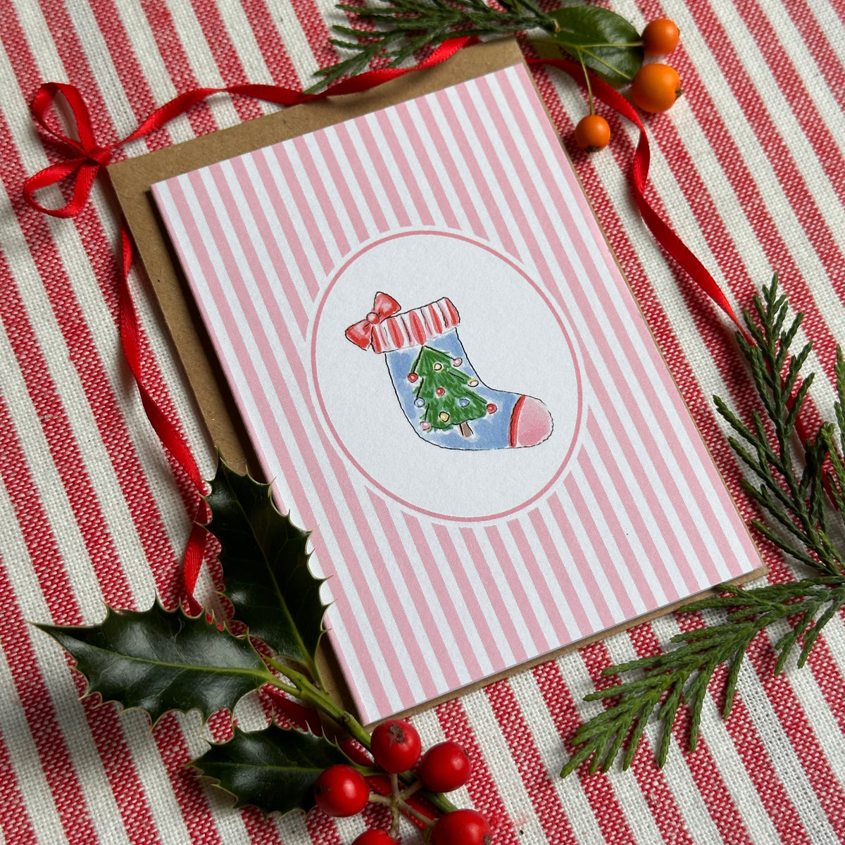 A Christmas-themed greeting card with pink and white vertical stripes and a central illustration of a stocking, styled on a red and white striped fabric with holly, pine branches, and red berries.