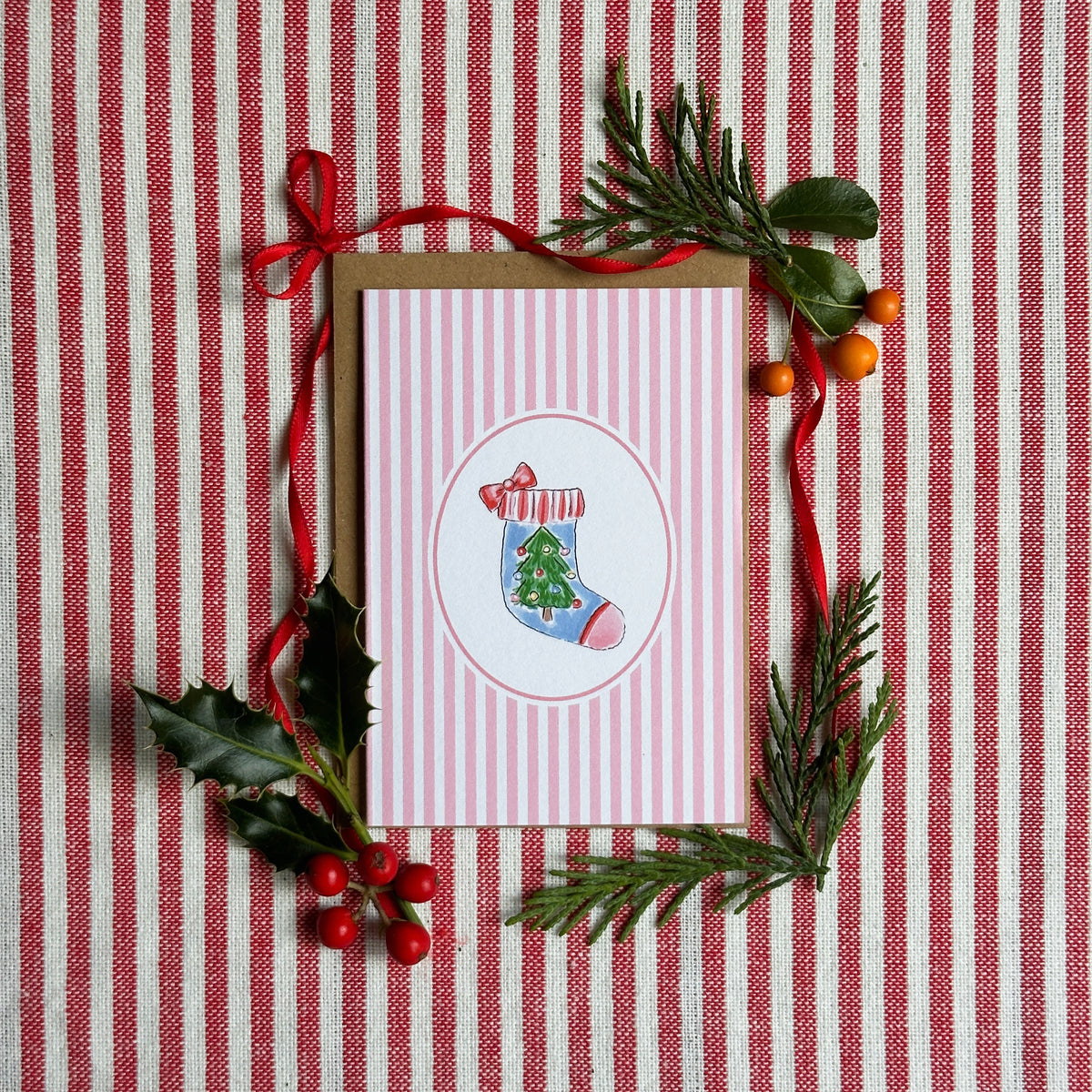 A Christmas-themed greeting card with pink and white vertical stripes, featuring an illustration of a blue stocking with a green Christmas tree. The card is displayed on a brown kraft envelope, surrounded by festive green pine branches, holly leaves, and red berries on a red and white striped fabric background.