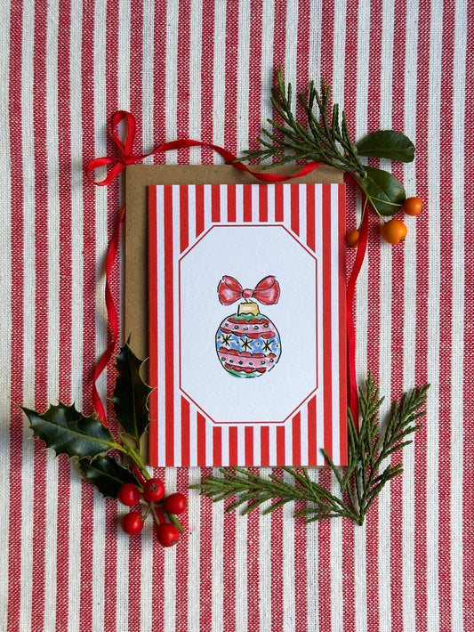A Christmas greeting card with red and white vertical stripes and an illustration of a decorated ornament, styled with a kraft envelope, red ribbon, holly leaves, pine branches, and berries on a red-and-white striped fabric background.