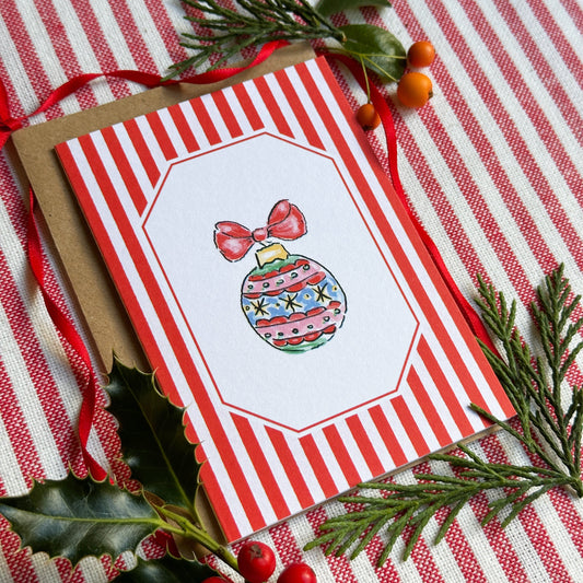 A Christmas greeting card with red and white stripes and an illustrated ornament, styled on a red-and-white striped fabric with holly, pine branches, and berries.