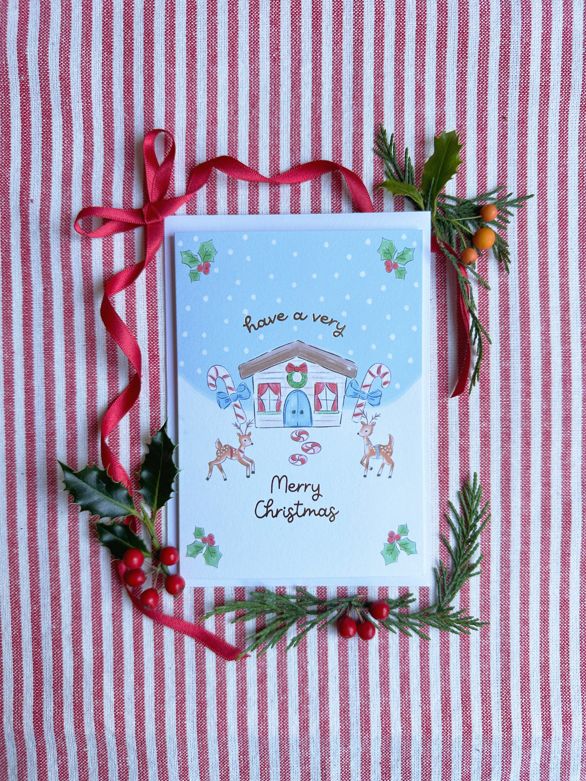 A Christmas greeting card with a winter house illustration and the words 'Have a very Merry Christmas,' styled on a red-and-white striped fabric background with a red ribbon and greenery accents.