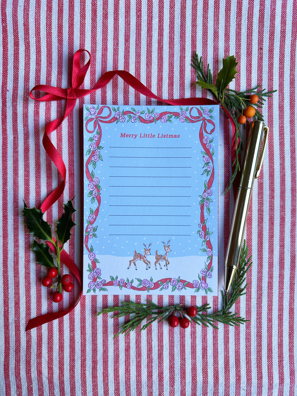 A6 Tear Off sheet Notepad with red ribbon and floral border blue background and two deer. Top of Notepad reads 'Merry Little Listmas". Backdrop is styled on a red and white striped fabric, gold pen, red ribbon and greenery and berries.