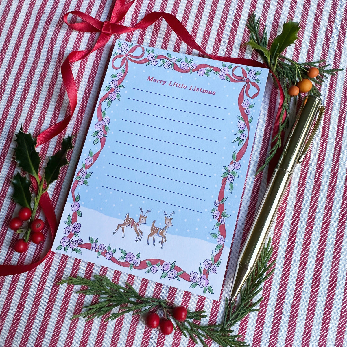 A6 Tear Off sheet Notepad with red ribbon and floral border blue background and two deer. Top of Notepad reads 'Merry Little Listmas". Backdrop is styled on a red and white striped fabric, gold pen, red ribbon and greenery and berries.