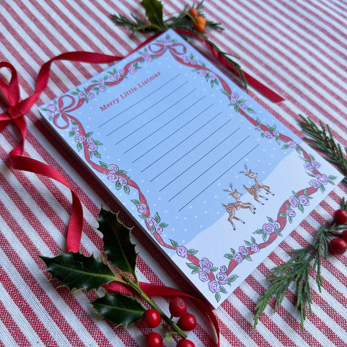 A6 Tear Off sheet Notepad with red ribbon and floral border blue background and two deer. Top of Notepad reads 'Merry Little Listmas". Backdrop is styled on a red and white striped fabric, red ribbon and greenery and berries.