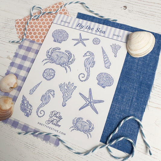 A sheet of decorative stickers featuring nautical designs like crabs, starfish, seahorses, shells, and coral in blue line art, styled on a white wood surface with blue-and-white twine, a gingham cloth, patterned paper, and seashells.