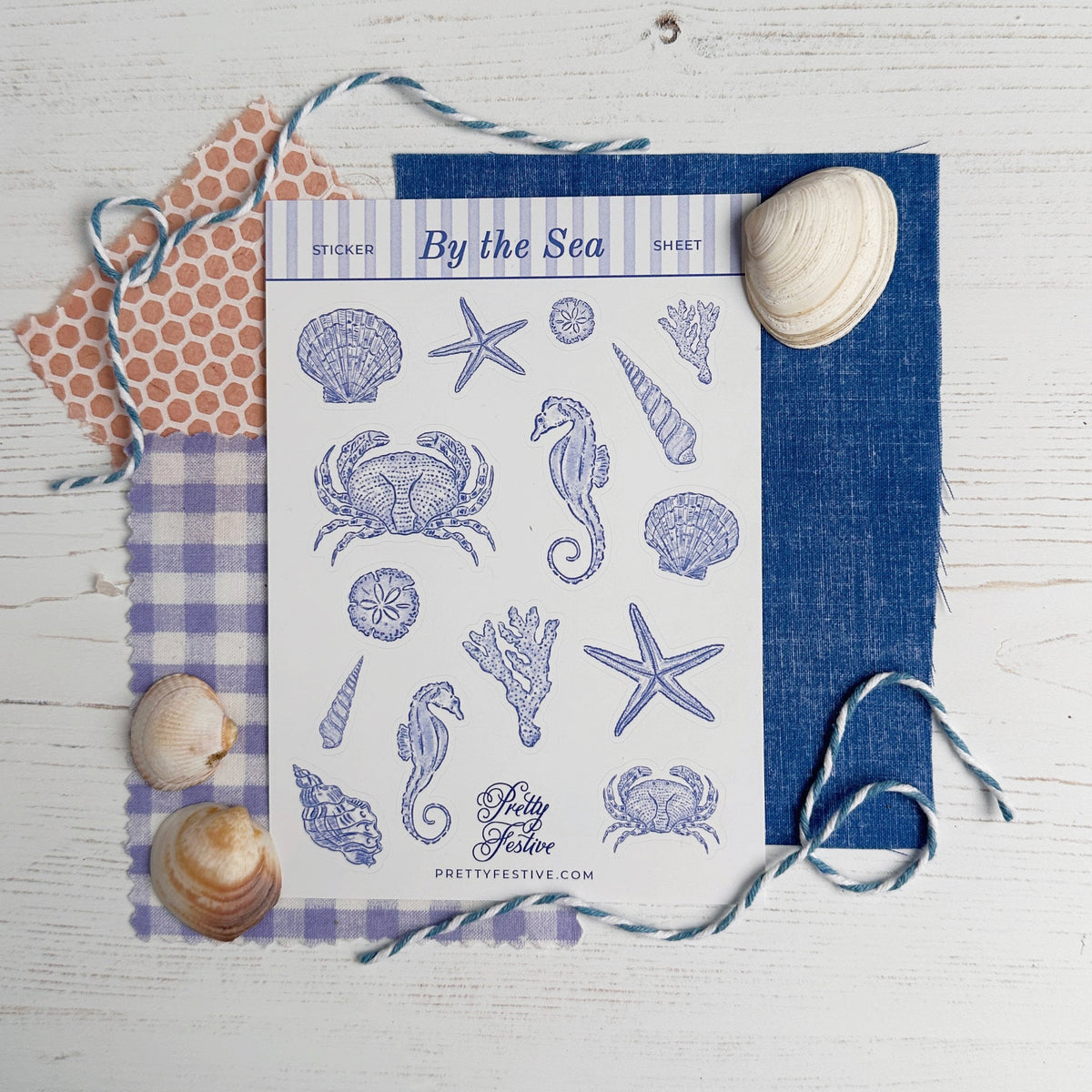A flat-lay product photo of a 'By the Sea' sheet of craft stickers on a white wood background, styled with blue and purple checkered fabric, a blue denim card, and seashells.