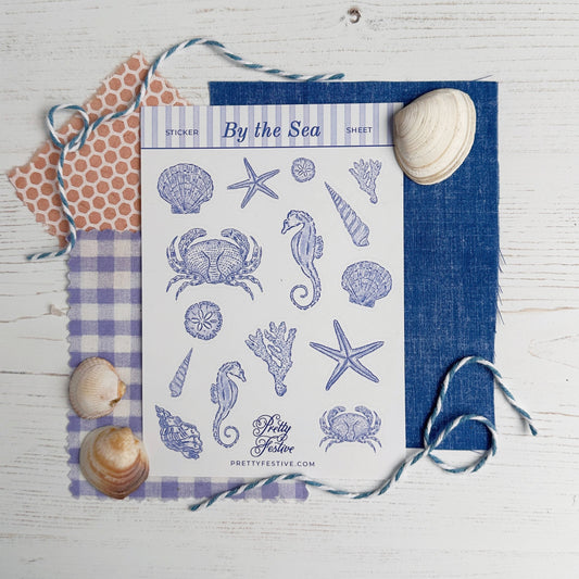 A flat-lay product photo of a 'By the Sea' sheet of craft stickers on a white wood background, styled with blue and purple checkered fabric, a blue denim card, and seashells.