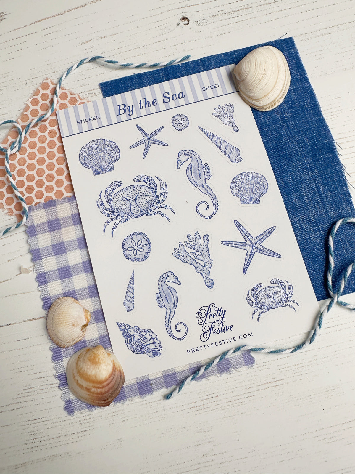 A sheet of nautical-themed Stickers titled 'By the Sea' featuring blue illustrations of crabs, starfish, seahorses, and shells, styled on a white wood background with seashells and patterned paper.