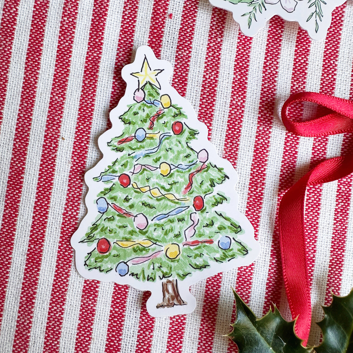 Vinyl die cut sticker of illustrated christmas tree. Background is red and white striped fabric with red ribbon greenery and berries.