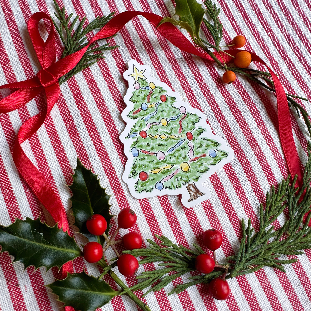 Vinyl die cut sticker of illustrated christmas tree. Background is red and white striped fabric with red ribbon greenery and berries.