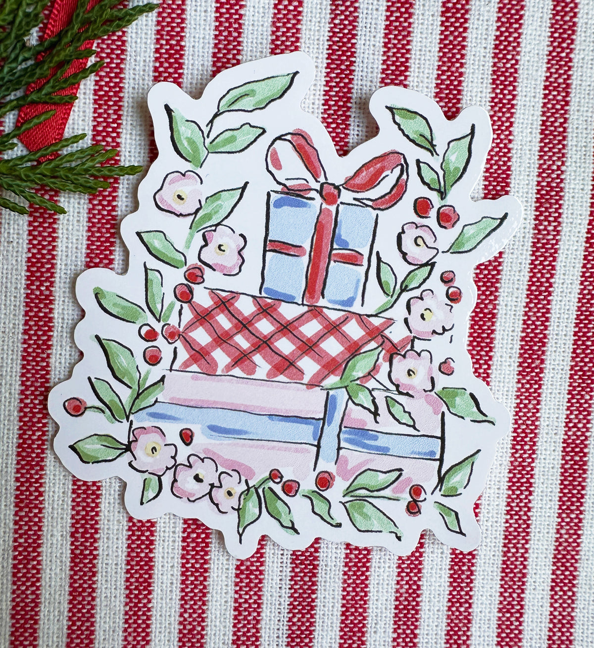 A die-cut sticker featuring a hand-painted style illustration of stacked gift boxes surrounded by green leaves and pink flowers, placed on a red and white striped fabric background.