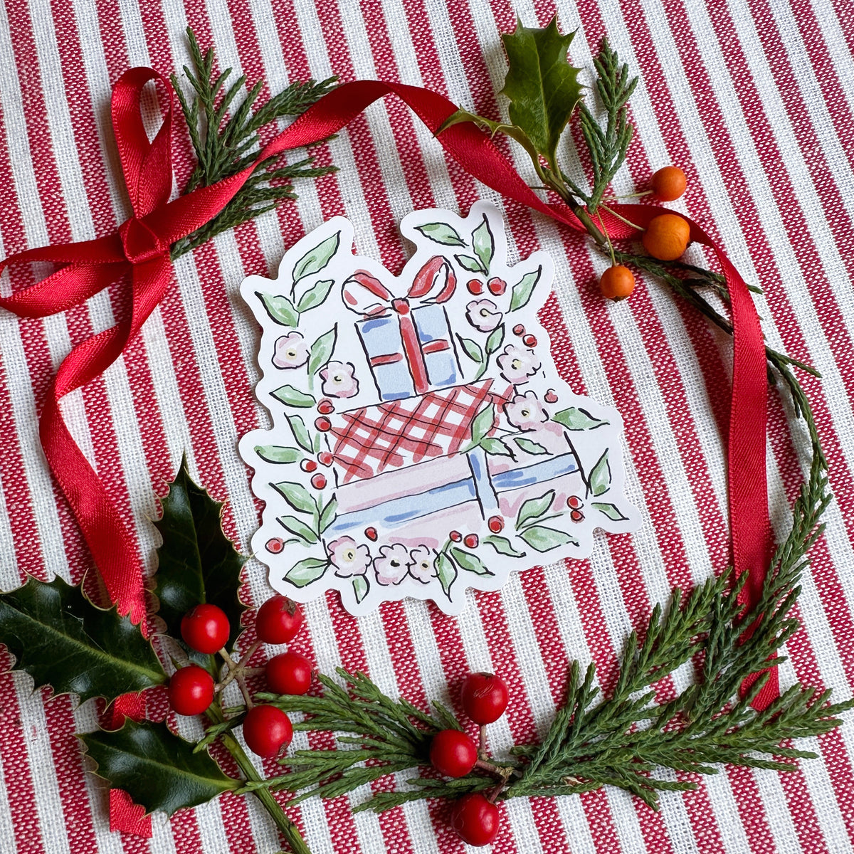 Vinyl die cut sticker of illustrated christmas gift stack surrounded by foliage and flowers. The background is red and white striped fabric with red ribbon greenery and berries.