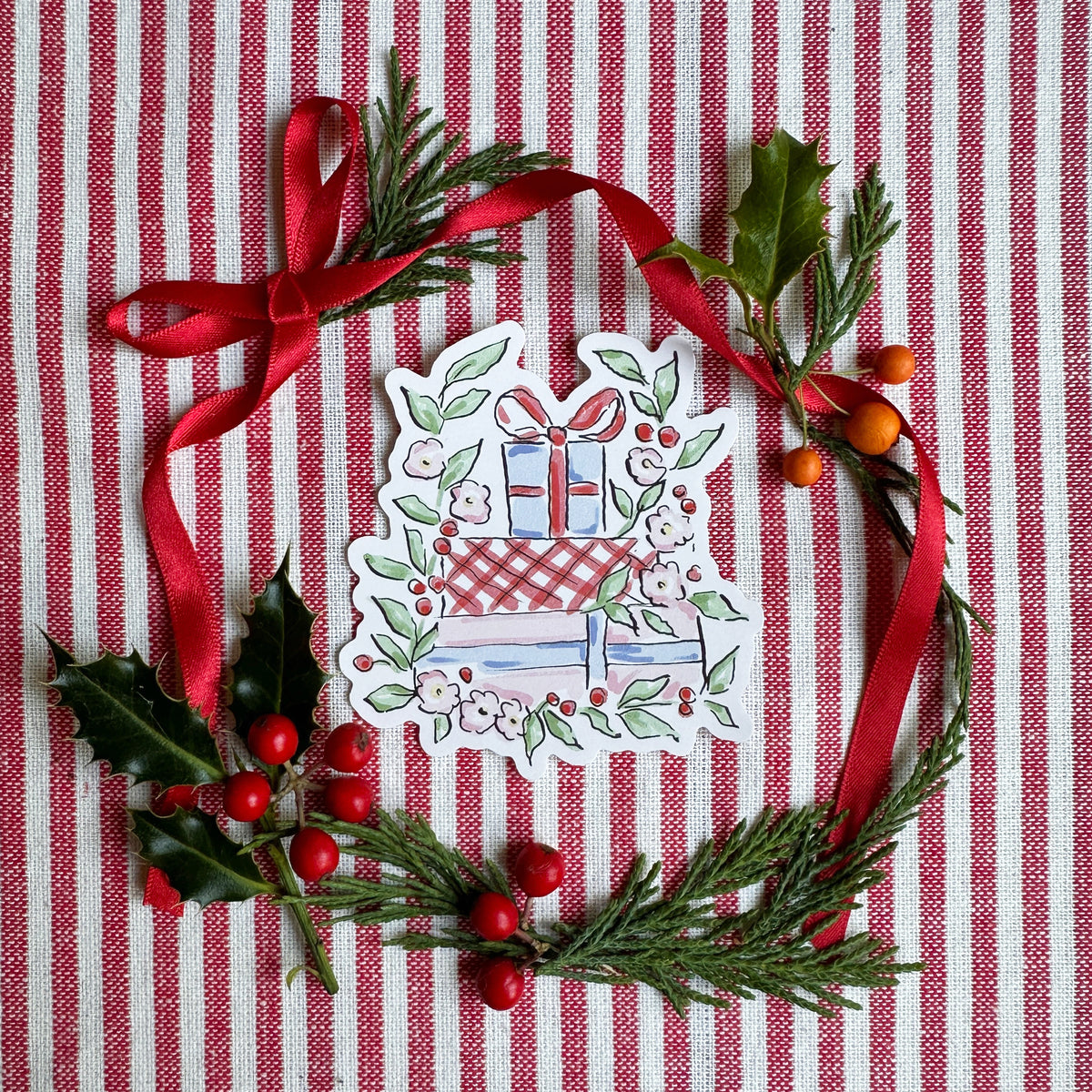 A die-cut sticker featuring a Christmas gift box with wrapped presents, surrounded by a hand-drawn wreath of green pine branches, holly leaves, red berries, and a red ribbon, all arranged on a red and white striped fabric background.