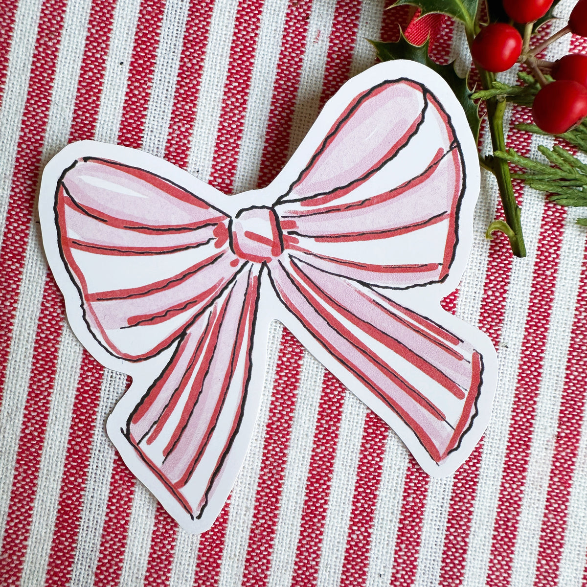 Vinyl die cut sticker of illustrated pink red and white bow. Background is red and white striped fabric with red ribbon greenery and berries.