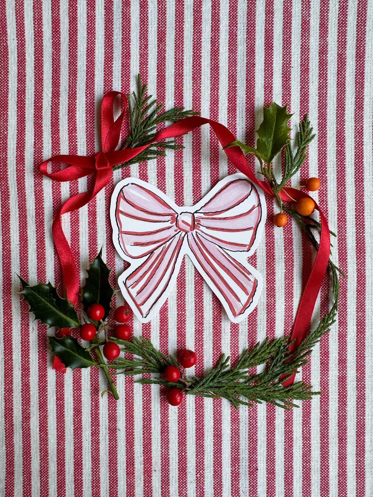 Vinyl die cut sticker of illustrated pink red and white bow. Background is red and white striped fabric with red ribbon greenery and berries.