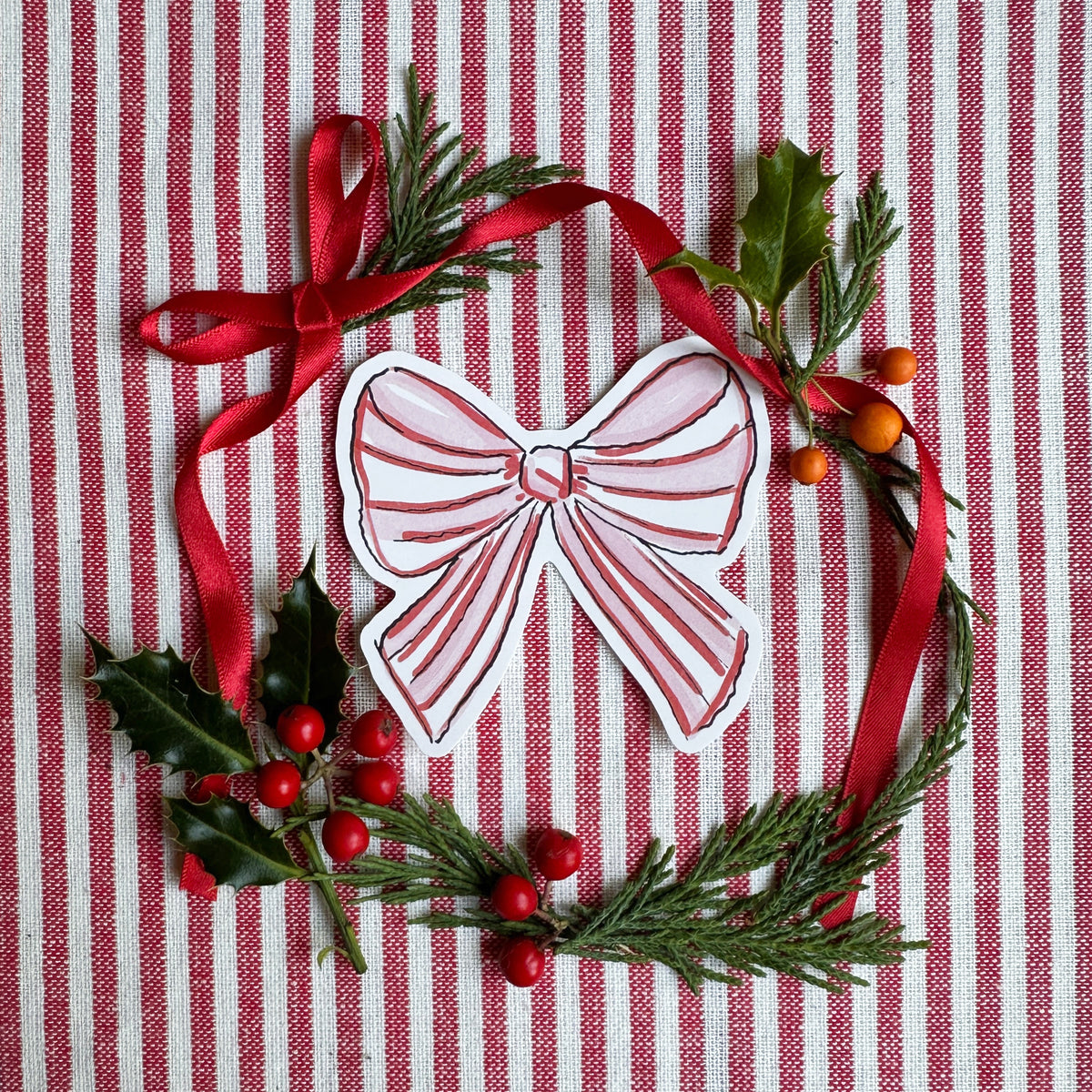 Vinyl die cut sticker of illustrated pink red and white bow. Background is red and white striped fabric with red ribbon greenery and berries.