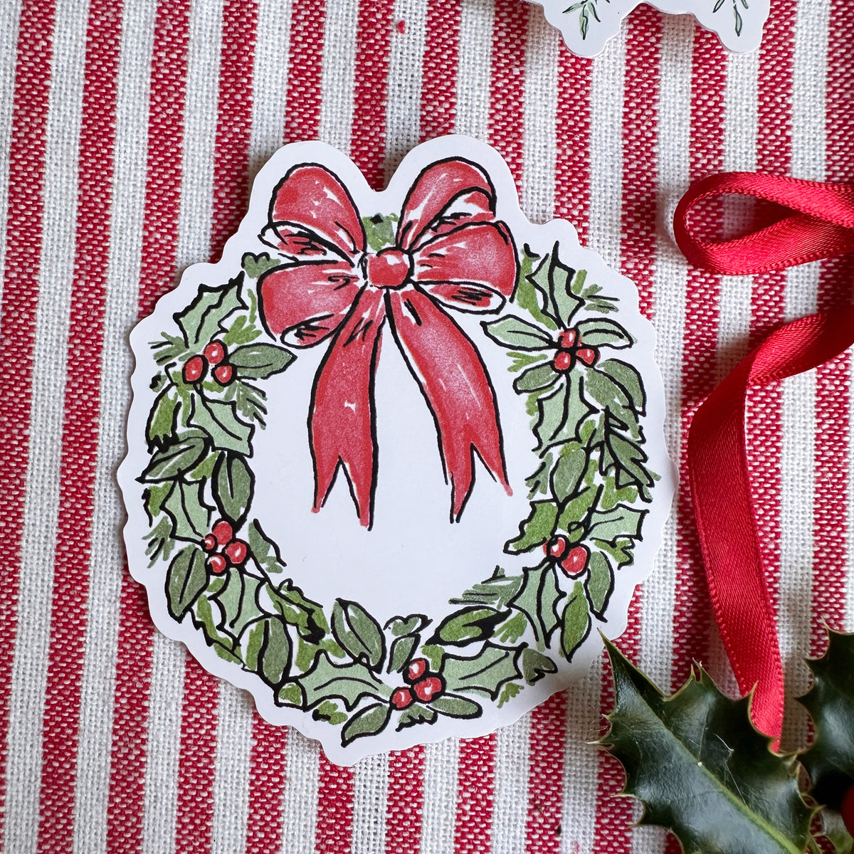 Vinyl die cut sticker of illustrated christmas wreath with red bow. Background is red and white striped fabric with red ribbon greenery and berries.