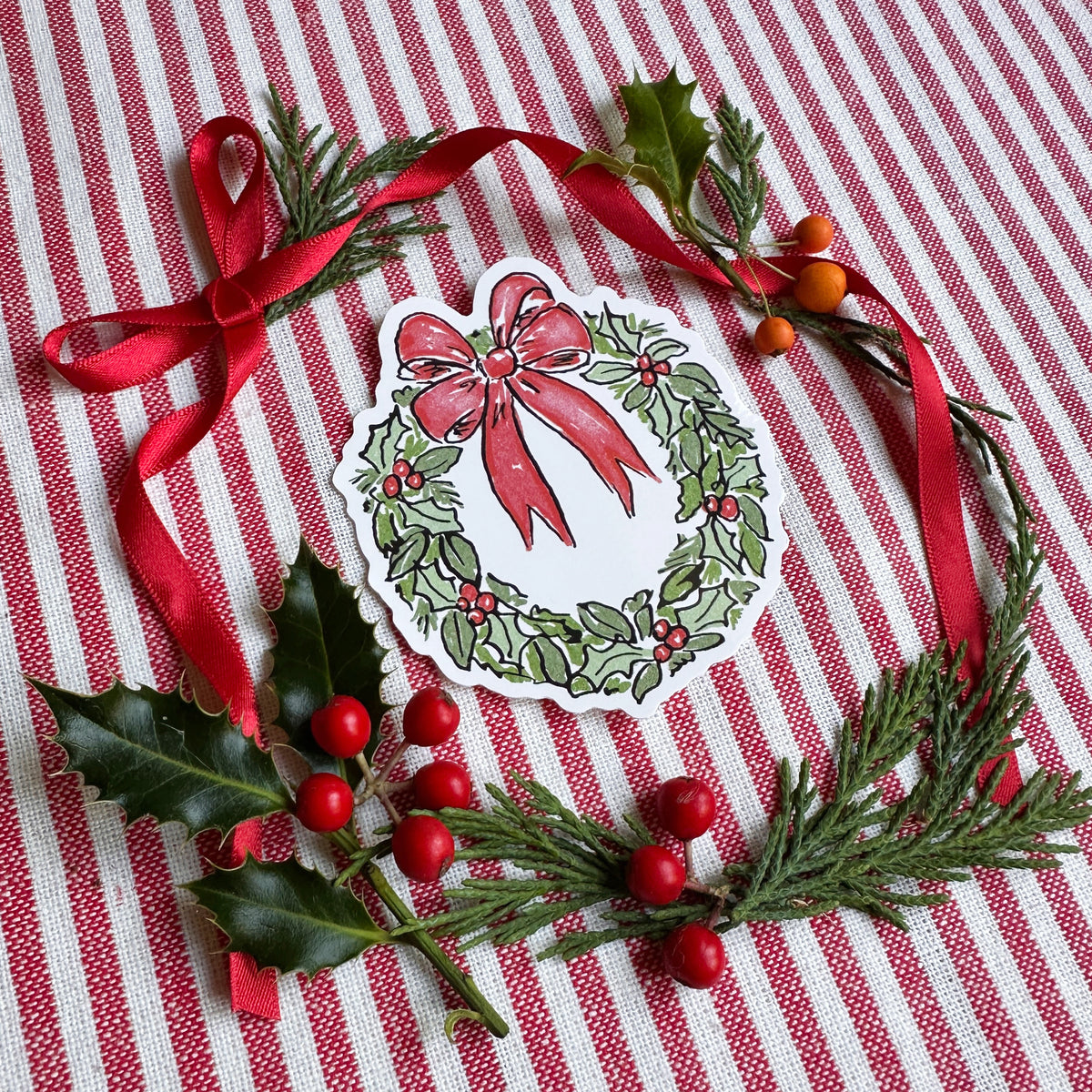 Vinyl die cut sticker of illustrated christmas wreath with red bow. Background is red and white striped fabric with red ribbon greenery and berries.