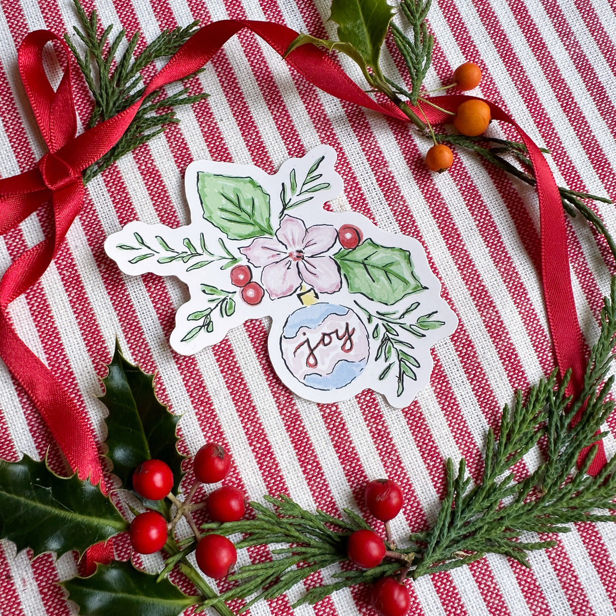 Vinyl die cut sticker of illustrated christmas ornament with foliage, berries and flowers. The background is red and white striped fabric with red ribbon greenery and berries.