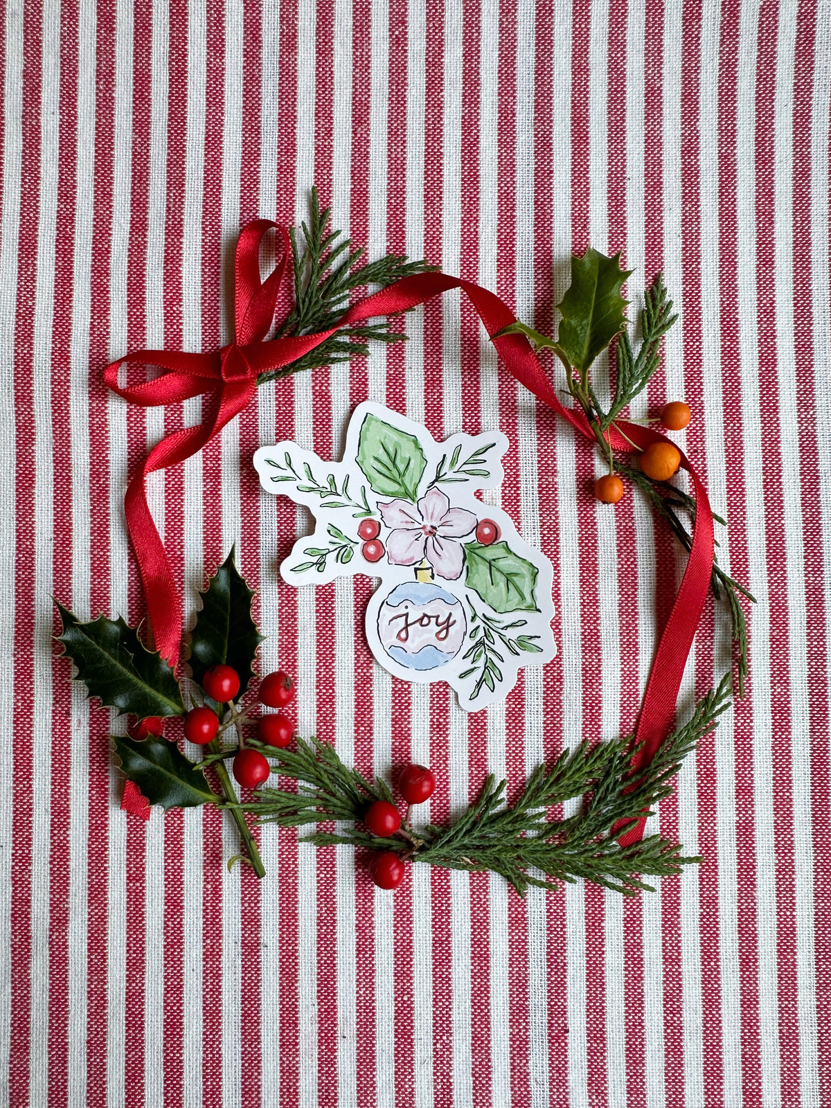 Vinyl die cut sticker of illustrated christmas ornament with foliage, berries and flowers. The background is red and white striped fabric with red ribbon greenery and berries.