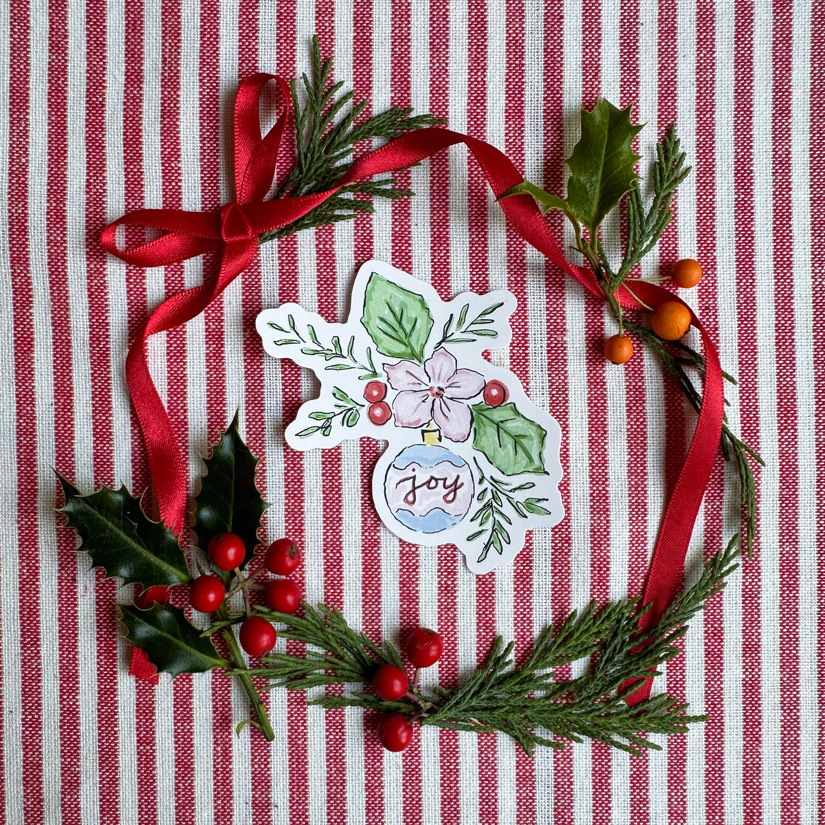 Vinyl die cut sticker of illustrated christmas ornament with foliage, berries and flowers. The background is red and white striped fabric with red ribbon greenery and berries.