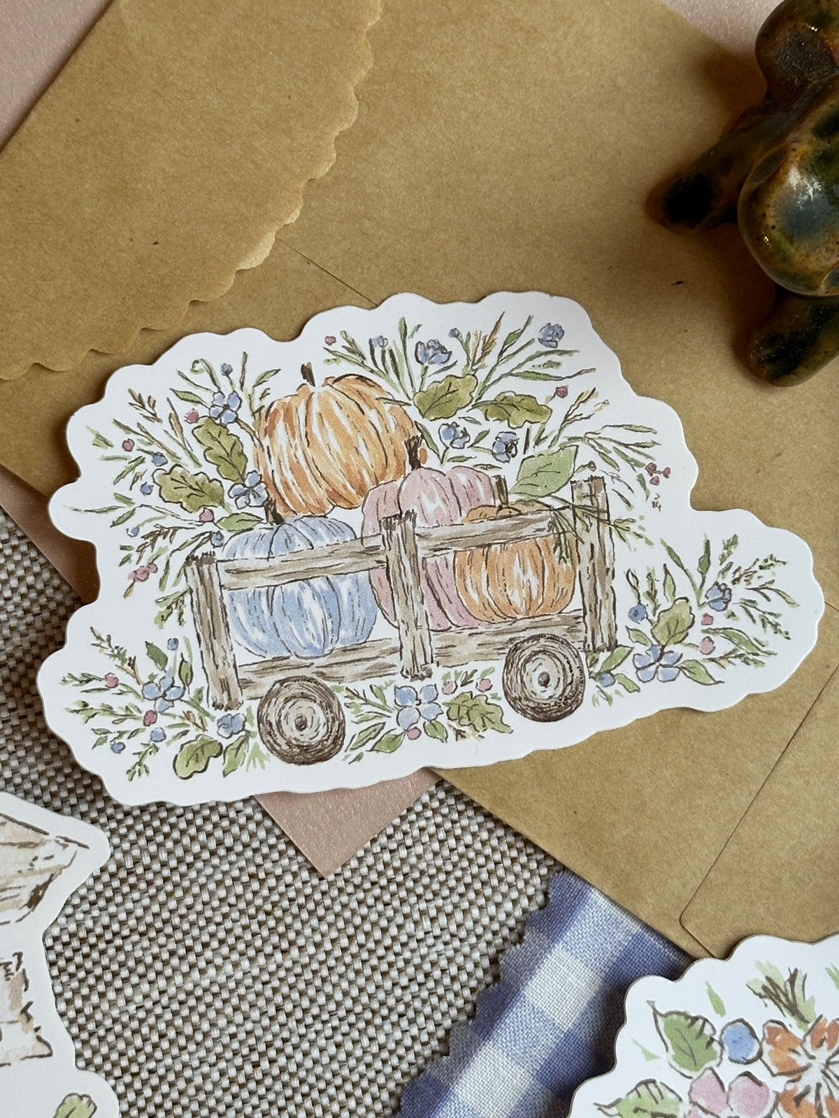 A die-cut sticker featuring a watercolor illustration of pumpkins in a wooden wagon surrounded by autumn leaves and flowers, displayed on a kraft paper envelope and textured fabric surface.