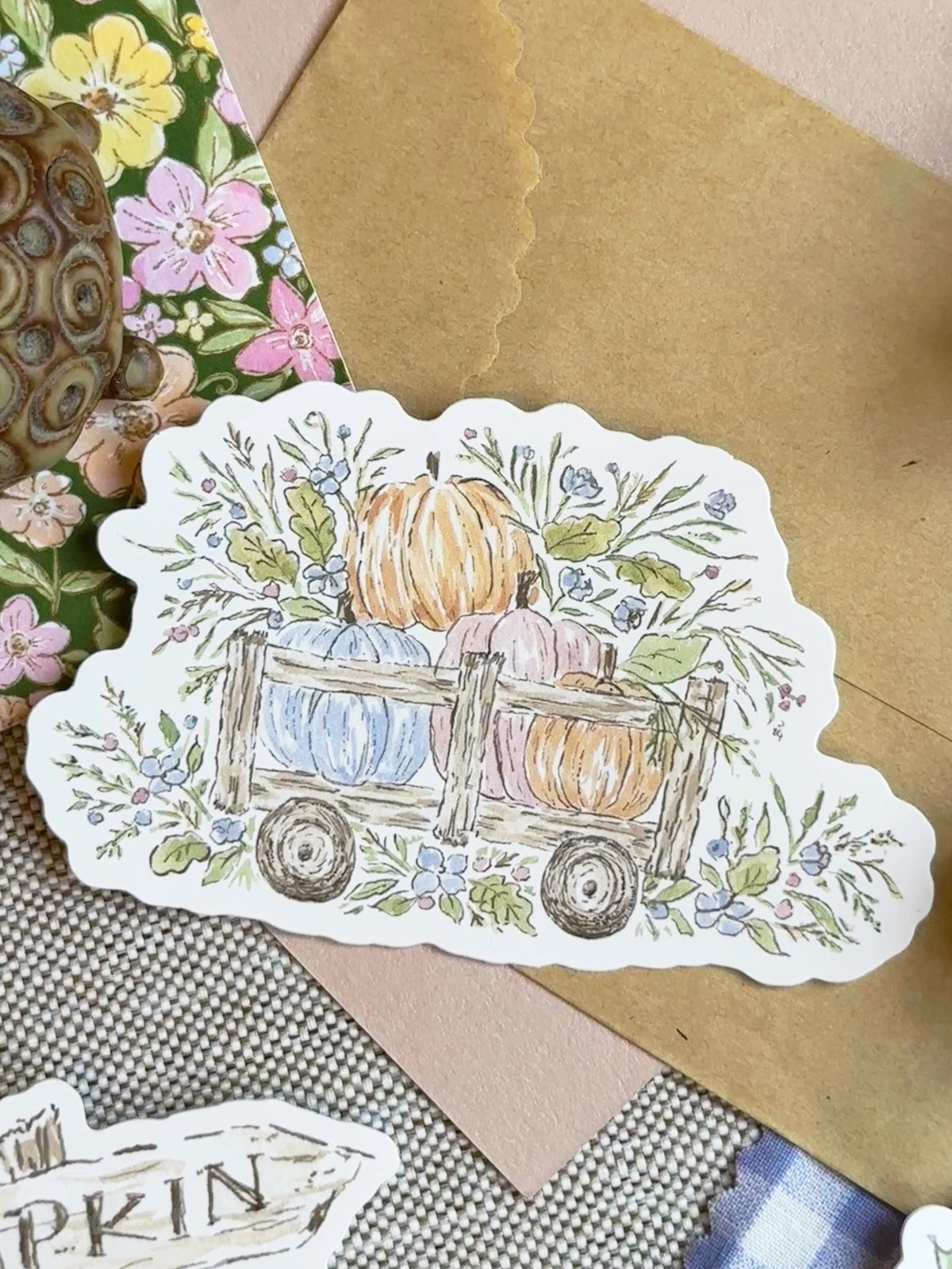 A scalloped die-cut sticker featuring a hand-drawn autumn illustration of pumpkins in a wooden wagon surrounded by flowers, displayed on a kraft paper envelope and fabric background.
